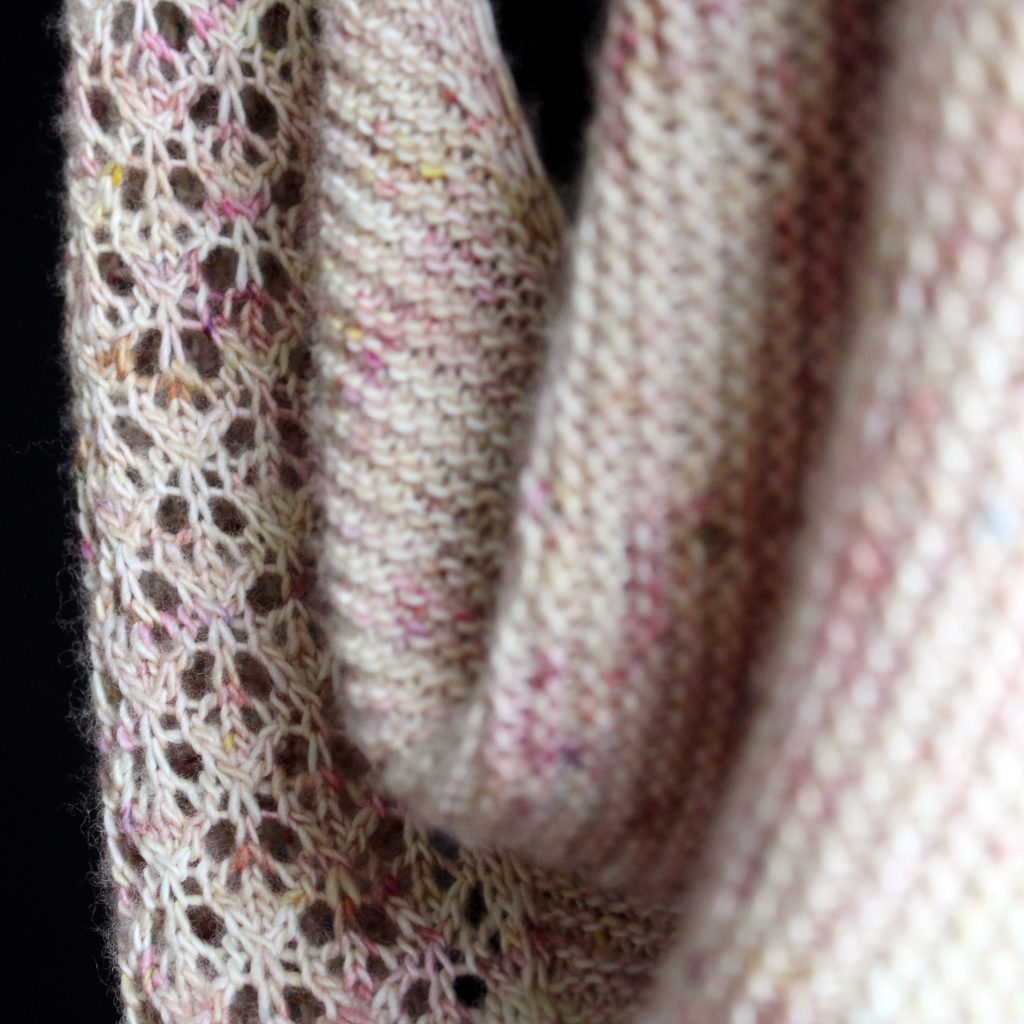 A close up on a the lace border and garter stitch body of a shawl knit in pale yellow speckled yarn