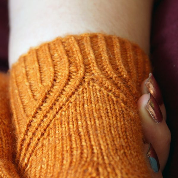 The cuff of fingerless mitts in orange yarn with a twisted rib pattern that splits to either side around a stockinette section.