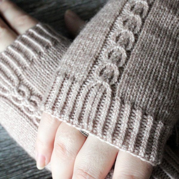 A close up on the cuffs of a pair of fingerless mitts with a twisted cable pattern, knit in a pale tan yarn